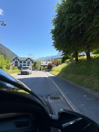 The image shows a view from inside a vehicle driving on a road in Les Houches, France. The road is paved, with a concrete curb and a storm drain visible. On the right side of the road, there is a grassy embankment with trees casting shadows. In the distance, there are houses with dark roofs, a crane, and mountains under a clear blue sky. A white van is driving in the opposite direction on the road. The interior of the vehicle, including the dashboard and side mirror, is partially visible in the foreground.