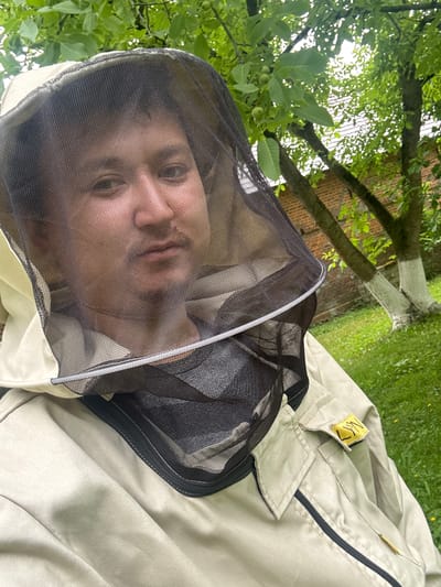 A person, seen from a close-up perspective, wears a beekeeping suit and veil. The veil's mesh partially obscures their face, but their eyes, nose, and mouth are visible. The suit is a light tan color, with a black zipper visible on the front. A small yellow tag with black writing is attached to the chest area of the suit. The background is blurred, showing green grass, trees with green leaves, and a hint of a brick wall. The lighting suggests it is daytime, possibly overcast or shaded by the trees. The overall impression is of someone dressed and ready for beekeeping, perhaps in a garden or natural setting. The image is oriented sideways, with the person's head appearing towards the bottom of the frame. The setting appears to be outdoors in a natural environment.