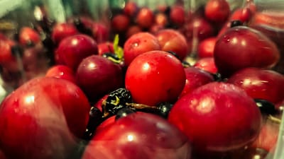 The image is a close-up, macro shot of a pile of ripe, red plums. The plums are glistening with what appears to be water droplets, suggesting they have been recently washed or are very fresh. Some dark berries, possibly blueberries or blackcurrants, are interspersed among the plums. The lighting creates bright highlights on the smooth surfaces of the fruit, giving them a glossy appearance. The background is softly blurred, with hints of green and other colors, but the focus remains squarely on the plums. There are no people or discernible elements of a specific location like the city of Troițcoe, Moldova, in the image. The scene is entirely focused on the texture and vibrant color of the fruit, presented in a shallow depth of field.