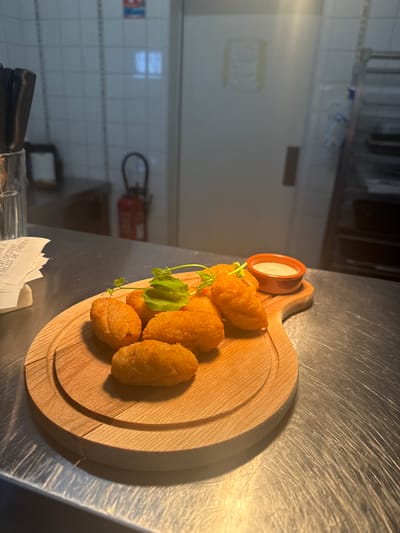 A close-up shot shows a wooden platter with several golden-brown croquettes, garnished with a sprig of green herbs. A small orange dish filled with a white dipping sauce sits next to the croquettes. The platter rests on a metallic surface, likely a countertop. In the background, a tiled wall, a fire extinguisher, and a metal shelving unit are visible. A crumpled receipt is partially visible on the left side of the image. The scene appears to be in a restaurant kitchen or service area in Rivesaltes, France. There are no people visible in the image.