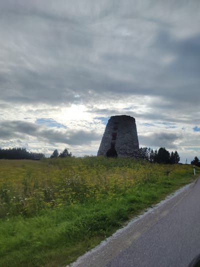 A stone windmill ruin stands on a grassy hill under a cloudy sky in Edise, Estonia. The windmill is conical in shape with dark openings and appears to be made of weathered gray stones. In front of the windmill, a field of green grass and yellow wildflowers slopes down towards a grey asphalt road that curves to the right of the frame. A line of trees is visible behind the windmill. The sky is filled with dramatic, dark gray clouds with patches of blue sky and sunlight breaking through.