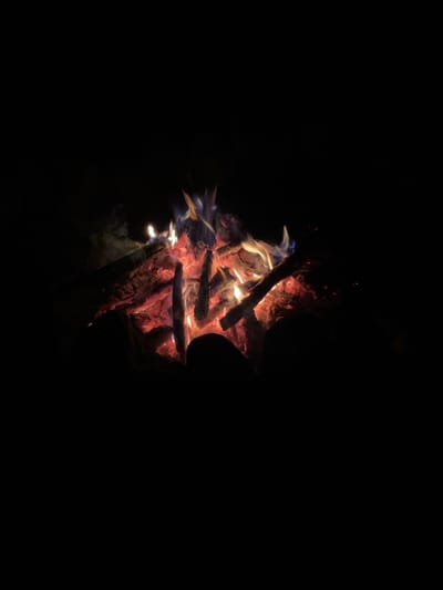 A campfire burns brightly in the darkness. Logs are stacked and smoldering with red-hot embers, producing vibrant orange and yellow flames that lick upwards. Wisps of smoke curl from the fire, disappearing into the blackness. The surrounding area is obscured by shadow, with only faint hints of dark, indistinct shapes suggesting rocks or the ground. The scene evokes a sense of warmth and isolation, typical of an outdoor setting at night.