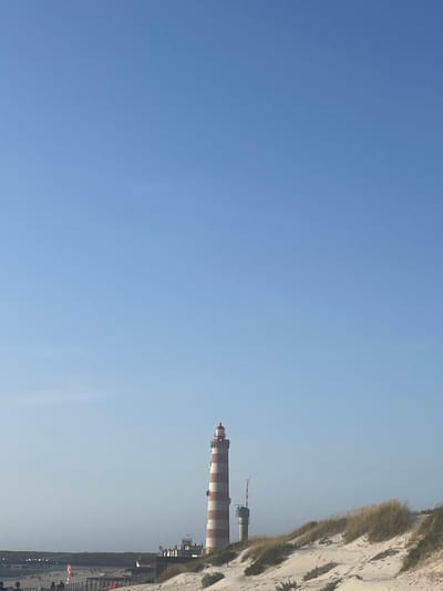 The image shows the Farol de Aveiro, a lighthouse located in Gafanha da Nazaré, Portugal. The lighthouse is tall and striped with red and white bands. To the right of the lighthouse, there is a smaller tower with an antenna. The scene is set on a sandy beach with dunes covered in dry grass and some sparse vegetation. In the background, there are some buildings and a wooden boardwalk, with a few people visible on or near the boardwalk. The sky is clear and blue.