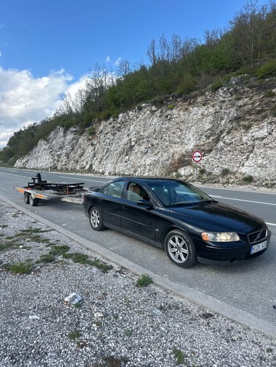 A black Volvo sedan with the license plate KOV 410 is parked on the right shoulder of a two-lane road next to a rocky cliff face. The car is attached to a small, flatbed tow dolly. A round, red and white traffic sign, indicating a prohibition, is posted on the cliff side of the road. The location appears to be outdoors in a mountainous area in Očinići, Montenegro, as evidenced by the landscape. Debris and small vegetation are visible on the gravel shoulder. The sky is partly cloudy.