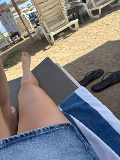 A person relaxes on a lounge chair. A striped towel lines the seat. Black flip-flops rest nearby on the sandy ground. The individual wears a denim skirt, and their legs are tanned. Yellow polish adorns the toenails. Several similar lounge chairs and beach umbrellas are in the background.