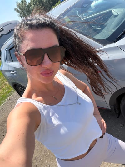A woman is captured in a selfie outdoors, with a grey SUV partially visible in the background. She is wearing a white cropped tank top and light-colored leggings. Her dark hair is pulled back in a ponytail and is being blown by the wind. She has large sunglasses on and is wearing a delicate silver necklace. The ground appears to be gravel or dirt, suggesting a rural or suburban setting. The sky is clear and bright, indicating a sunny day. No specific landmarks of Пригородный, Russia are identifiable in the image.