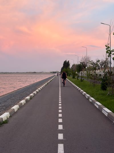 The image depicts a paved path, likely a bike path or promenade, bisected by a dashed white line. A person is riding a bicycle along the path, moving away from the viewer. A row of street lights lines the path on the left, with green vegetation and trees behind them. On the opposite side of the path is a low wall or embankment. Beyond the wall is a body of water. The sky is visible in the background, displaying a soft, pastel-colored sunset. The location is in Kuleshovka, Russia.