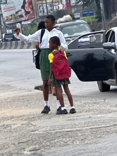 Two school children in uniform stand on a sidewalk next to a car. One wears a red backpack over a yellow shirt and a green and plaid skirt, while the other wears a white button-down shirt, dark pants, and carries a bag on their shoulder. Both wear white socks and dark shoes. A car door is open next to them. A billboard is visible behind them.