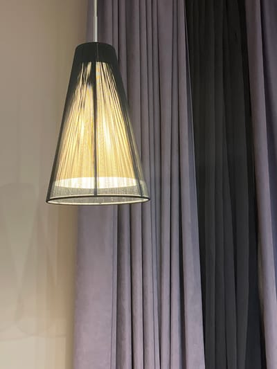 A modern cone-shaped pendant light fixture hangs in front of dark grey and light grey vertical curtains. The light fixture has a black frame and a translucent shade made of thin, vertical strands that emit a warm yellow light. The wall to the left of the fixture is a plain, light beige color. The image does not provide any information about the city of Otyakovo, Russia, or any people or actions.