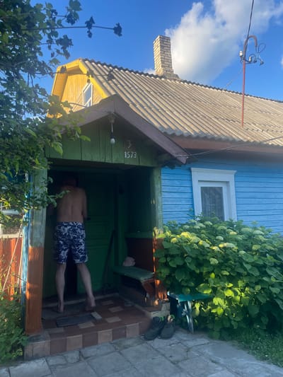 A man wearing patterned shorts and no shirt stands with his back to the camera, entering the doorway of a rustic blue-and-yellow house. The house has a grey, corrugated roof and a brick chimney. In front of the house, a dense green bush with white flowers is in full bloom. To the left of the entrance, a wooden structure is partially visible, painted orange and green. On the ground near the entrance, a pair of dark boots rests. The scene appears to be in a rural setting, possibly the village of Блужскі сельскі Савет, Belarus, given the architectural style and natural surroundings.