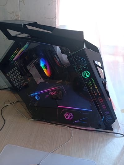 A black computer tower with a transparent side panel sits on a wooden floor. The computer has illuminated RGB lighting on its components, including the CPU cooler and graphics card. The tower features a distinctive angular design with cutouts and decorative patterns. Cables are visible connecting to the computer. In the background, a sheer white curtain is partially visible, and the wall behind it appears textured. The image is an interior shot, taken from a slightly elevated angle, with the computer positioned in the foreground. There are no people or animals present in the scene. The location is identified as Khavast, Uzbekistan.