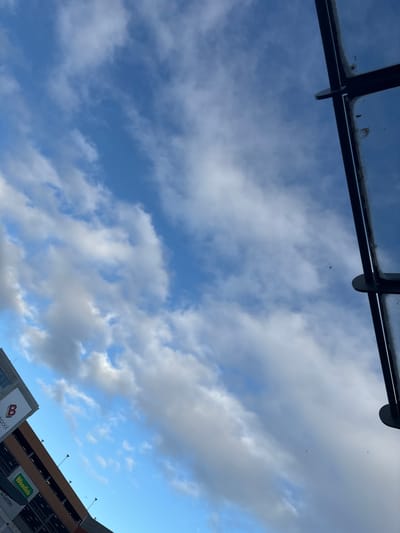 The image captures a view of a bright blue sky with scattered white clouds, taken from a low angle. In the upper right portion of the frame, a dark, angular structure, possibly part of a building or canopy, intrudes into the scene. Along the left side of the image, a multi-level building is visible, identified by signage including a large "B" logo and the text "Woodies". This building appears to be a car park, with multiple levels of open-air parking visible. The scene suggests an outdoor setting in a built-up area, with the sky dominating the upper portion of the composition. The building with the "Woodies" signage is located in Cork, Ireland.