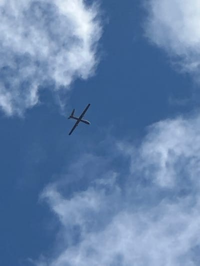 An unmanned aerial vehicle (UAV) is depicted against a bright blue sky with scattered white cumulus clouds. The dark-colored UAV, visible from a low angle, features a slender fuselage, straight wings, and a T-tail, indicating it is in flight. No individuals or other discernible objects are present in the frame. The scene is located in the city of Beit Yitshak Sha'ar Hefer, Israel.
