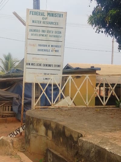 The image shows a sign for a project by the Federal Ministry of Water Resources. The Anambra-Imo River Basin Development Authority is undertaking the project, which involves the provision of a motorized borehole at NRI. Hon. Mrs. Uche Ekwunife (IYOM) is listed as having attracted the project. The contractor is Solar Product Limited. The sign is old, weathered, and mounted on posts next to a low wall. The setting appears to be a rural area. A partially visible sign reading "BUY IT" is on the lower left.