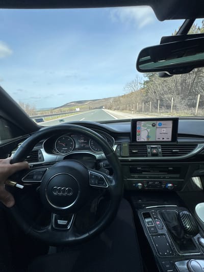The scene depicts the interior of an Audi vehicle from the driver's perspective, positioned on a multi-lane highway near Kipra, Bulgaria.
**Environment (Exterior):** The vehicle is traveling on a paved highway with at least two lanes visible in the direction of travel, marked with white lines. Guardrails line both sides of the road. The immediate roadside features dry grass and sparse, bare deciduous trees, indicative of late winter or early spring. Hilly terrain flanks the highway, extending into the distance. A body of water, likely a sea or large lake, is visible on the horizon to the left. The sky is predominantly clear blue with scattered white clouds. A white utility marker is visible on the right shoulder.
**Interior (Vehicle):** The vehicle is an Audi, identifiable by the four-ring logo on the steering wheel. The dashboard displays the instrument cluster, showing a speed of approximately 128 km/h both on the analog speedometer and digitally. An outside temperature of 25°C is also indicated on the digital display. The driver's left hand, with light skin tone, is visible gripping the lower-left spoke of the steering wheel. This hand is also holding a gold-tipped vaping device. An infotainment screen, centrally mounted on the dashboard, displays an Apple CarPlay interface with a navigation map (Apple Maps) showing the vehicle's position. The screen indicates "11:45 AM" and "12°C." Below the screen, climate control settings are visible, both set to 22.0°C. To the right of the driver, the center console features an automatic gear selector with a carbon fiber-like finish, along with control buttons for the parking brake and MMI system. A rearview mirror is mounted on the windshield. A rectangular, green device is clipped to an air vent to the left of the infotainment screen. The lower body of the driver, clad in dark trousers, is partially visible.
**Actions and State:** The vehicle is in motion at 128 km/h. The driver is operating the vehicle and simultaneously holding a vaping device. The navigation system is active.