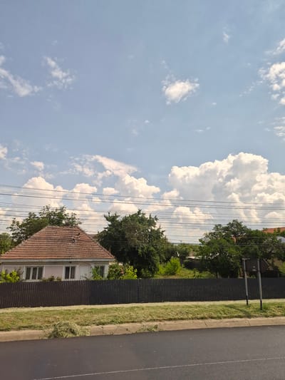 The image depicts a suburban street scene in Răcăciuni, Romania, on a partly cloudy day. In the foreground, a paved road runs horizontally across the bottom of the frame. To the right of the road, a strip of grass borders a concrete curb, with a pile of freshly cut grass clippings visible on the roadside. A dark, wooden slatted fence runs along the property line behind the grass. Beyond the fence, a modest, single-story house with a terracotta tile roof is visible, featuring several windows and a white door. Lush green trees and vegetation are present behind the house and along the fence line. Overhead, multiple black electrical wires are strung across the frame. The sky above is a pale blue with scattered white, puffy cumulus clouds. The overall impression is one of a quiet, residential area during daylight hours.