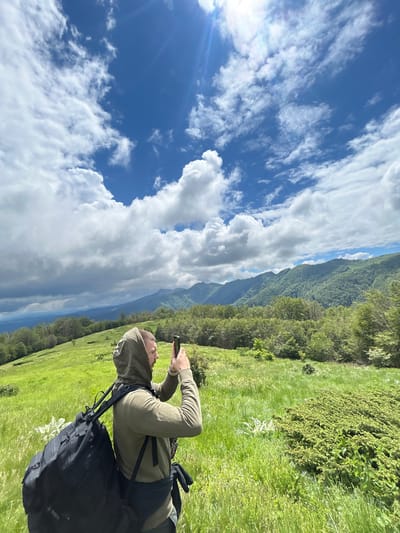 In Ribaritsa, Bulgaria, a man with a backpack stands in a grassy field, taking a picture with his phone. He is wearing a green hooded sweatshirt. The field slopes gently upwards towards a lush, forested mountain range under a partly cloudy blue sky. The sun shines brightly, casting a lens flare onto the image.