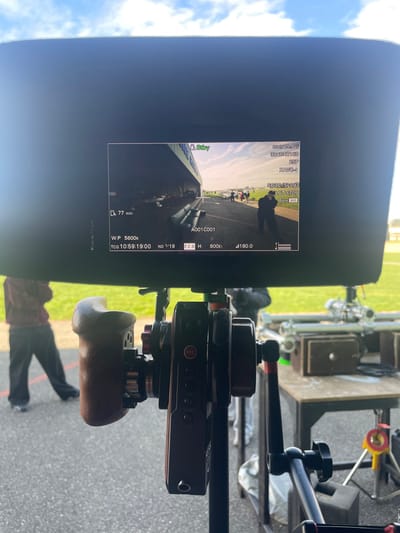An outdoor scene in Chonas-l'Amballan, France, depicts a film production setup. The central focus is a professional video monitor, framed by a black sun hood, displaying a live camera feed.
The monitor's screen shows technical data, including "3840x2160" resolution, "25P" frame rate, "XAVC-I" codec, "SG3C/SLOG3" color profile, "LUT 6709," "Base 800" ISO, "77 min" recording time, "A001C001" clip name, "W.P 5600k" white balance, "TCG 10:59:19:00" timecode, "ND 1/19" neutral density filter, "F2.8" aperture, "800Ei," and "180.0" degrees.
The live feed itself shows an expansive outdoor location under a bright, partly cloudy blue sky. A wide paved area, possibly asphalt, extends from under a large, open-sided structure with a green and white angled roof, reminiscent of a hangar or a covered grandstand. Beneath this structure, several dark, cylindrical objects are horizontally arranged. On the paved surface, numerous individuals are scattered, some walking or standing, with at least one person in dark attire visible in the mid-ground. Grassy fields are visible beyond the paved area, with more people in the distance.
In the immediate foreground, a professional cinema camera rig is visible, featuring a dark-colored body with a prominent red "REC" button and a brown wooden handgrip on the left side. Various gears, knobs, and support arms are part of the setup. To the far left, the lower body of an individual wearing dark pants and a maroon hoodie is visible, standing on the asphalt which has a red line marking. To the right of the camera rig, a metal table holds silver tubular equipment, possibly lighting stands or rigging components, and wooden boxes. A yellow equipment stand is partially visible beneath the table. The ground is primarily asphalt, transitioning to green grass in the background, under a sky with scattered white clouds.