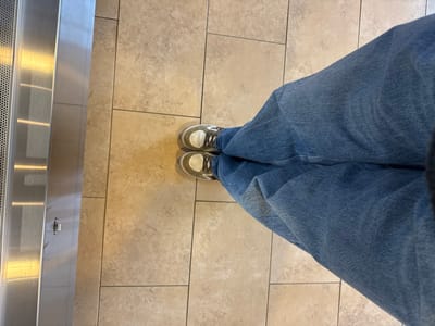 A first-person perspective shows a person standing on a tiled floor. They are wearing blue jeans and neutral-colored sneakers. To the left is a shiny, reflective surface, potentially metal, with linear designs or patterns. The floor tiles are light brown or beige and are arranged in a grid pattern. The individual's legs are the central focus.
