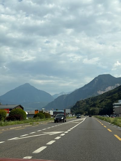 This media file captures a scenic view from a road in La Balmaz, Switzerland. The scene is dominated by imposing mountains covered in lush green forests, with the road winding alongside them. The sky is partly cloudy, suggesting it could be late morning or early afternoon.

On the right side of the frame, a dark asphalt road is clearly visible, marked with white dashed lines and yellow dashed lines, indicating it's a paved road with lanes. Several vehicles are parked along the side of the road, and some are driving on it, suggesting a normal day with traffic.

The left side of the image shows the dramatic mountainous landscape. The mountains are a deep blue-grey, contrasting with the green vegetation. In the foreground on the left, there's a glimpse of buildings with dark roofs and manicured green bushes, adding a touch of human settlement to the natural grandeur.

Visible text includes "UNICAR" and "JORGENSEN" on an orange sign, and what appears to be "FUJI" on a red sign. These suggest commercial activity or businesses in the vicinity. The overall mood is one of natural beauty and the coexistence of human infrastructure with the majestic Swiss landscape. The image appears to be taken from a moving vehicle due to the perspective and the slight blur in some elements.
