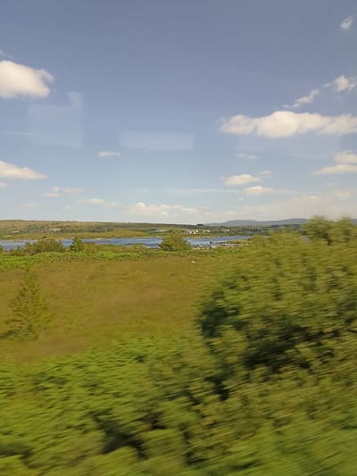 The image depicts a landscape scene, likely viewed from a moving vehicle given the motion blur in the foreground. The sky is a bright blue with scattered white clouds. Below the sky, there is a horizon line with rolling hills covered in green vegetation and some scattered buildings. A body of water, possibly a lake or bay, is visible in the mid-ground, with islands or peninsulas dotted within it. The foreground is dominated by a field of green grass and foliage, with some trees and shrubs. The vegetation in the immediate foreground is significantly blurred, suggesting rapid movement. The scene appears to be a rural or semi-rural setting, with natural elements like water, hills, and vegetation being prominent. There is no identifiable human presence or action in the image. The location is specified as Rampart, Ireland.
