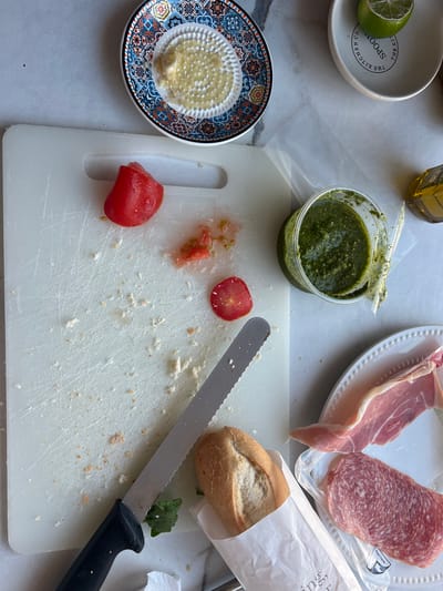 The image depicts a culinary scene, likely a kitchen counter, with ingredients for preparing a meal. A white cutting board is central, bearing remnants of chopped tomatoes and breadcrumbs, along with a serrated knife. To the upper left, a decorative plate holds grated garlic. A small bowl contains a segment of lime and is inscribed with "THE KITCHEN CO." In the upper right, a bottle of olive oil is partially visible. Adjacent to the cutting board is a container of green pesto. To the lower right, a white plate displays slices of cured meat, including prosciutto and salami. A partially unwrapped baguette sits next to the plate. The overall impression is of an unfinished preparation, possibly for sandwiches or a charcuterie board. The environment is a domestic setting, likely a kitchen. There are no discernible elements that would place this scene in a specific city or country, including Brighton, United Kingdom.