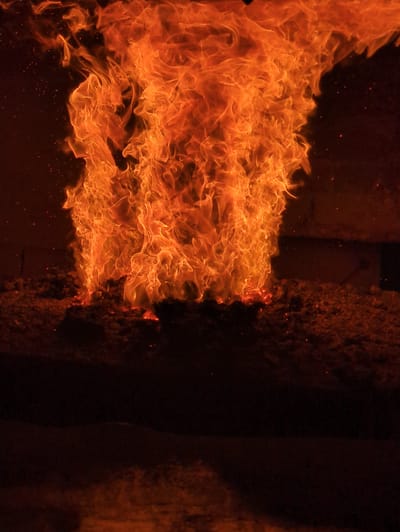 A large, vibrant flame engulfs the frame, originating from a pile of dark, granular material. The fire is an intense orange and yellow, with wisps and tendrils of flame reaching upwards and outwards, creating a dynamic, turbulent shape. Small red sparks are visible within and around the flames, suggesting intense heat. The foreground consists of the burning material, which appears to be spread out across a dark, uneven surface. In the background, a dark, solid structure is partially visible to the right, with some indistinct shapes suggesting a building or enclosure. The overall atmosphere is one of intense heat and combustion. This scene is located in Bolesławów, Poland.
