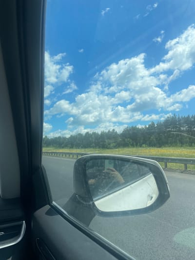 The image shows a view from inside a car, looking out the side window and into the side mirror. The sky is bright blue with scattered white clouds, suggesting a sunny day. The car appears to be traveling on a paved road, with a guardrail and a line of trees visible in the distance. The trees suggest a rural or suburban setting, possibly near a highway.
In the side mirror, a hand is visible holding a smartphone. The reflection in the mirror also shows the interior of the car and the road behind. The overall impression is of a journey, perhaps a road trip, on a clear and pleasant day. The scene is dynamic, captured from a moving vehicle, indicated by the slight blur and the perspective of the road. The image has a casual, everyday feel, capturing a moment of travel. There is no visible text within the image.
