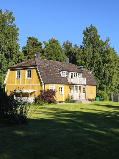 A bright yellow, two-story house with a dark, sloped roof stands in a grassy yard surrounded by lush green trees. The house has white trim around its numerous windows and a white-railed balcony on the upper floor. A small, white covered porch is visible on the first floor to the left of the entrance. The sky is a clear, vibrant blue, suggesting a sunny day. The sunlight casts shadows across the lawn, indicating it is likely midday or afternoon. The setting appears to be a suburban or rural residential area, as suggested by the spacious yard and abundant trees. There are no people visible in the image. A green garbage bin is present near the right side of the house, and a metal fence is visible in the far right background. The overall mood is peaceful and serene, with the bright colors and natural setting creating a cheerful atmosphere. No text is visible in the image.
