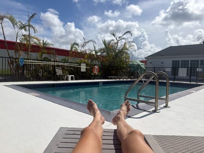 In Palm Bay, Florida, a person's legs and feet are visible from a first-person perspective as they recline on a lounge chair by a swimming pool. The pool is rectangular with a blue tiled edge and a depth marker indicating "4 FT." Palm trees and tropical foliage line the perimeter of the pool area, which is enclosed by a black metal fence. Beyond the fence, a building with a white exterior and a dark roof is visible, along with a red-colored building in the distance. The sky is partly cloudy with large white cumulus clouds against a bright blue background. A blue pool skimmer hangs on the fence, and a life preserver with a red ring is also visible.