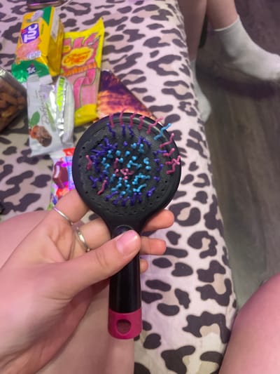 A close-up, eye-level shot shows a person's hand holding a black hairbrush with multi-colored bristles. The hand is positioned in the foreground, with fingers adorned with silver rings. The hairbrush is dark with purple, pink, and blue bristles. In the background, out of focus, is a surface covered with a leopard print fabric. To the left of the hairbrush, several candy wrappers are visible, including a yellow "TUC" cracker package and a pink "Chupa Chups" lollipop package. To the right of the hairbrush, a person's legs are visible, wearing grey socks and leaning against the leopard print surface. The overall lighting suggests an indoor setting. The image does not provide any information about the location in the city of Shcheglovo, Russia.