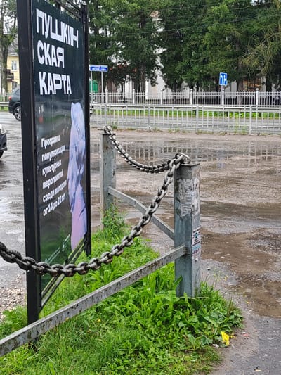 The image depicts a street scene in Vorsma, Russia, on a rainy day. A large black billboard stands on the left, displaying text in Russian and an image of a sculpture. The billboard reads "ПУШКИНСКАЯ КАРТА" (Pushkin Card) and further text describes a program for popularizing cultural events among young people aged 14 to 22. To the right of the billboard, a metal fence with thick chains is visible, separating the sidewalk from the street. The street is wet and reflects the overcast sky, indicating recent rain. Trees with green foliage line the background, and a few buildings can be seen in the distance. A blue road sign is also present in the background, indicating traffic direction. The overall impression is one of a typical urban environment during inclement weather.