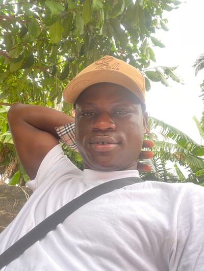A medium close-up outdoor photograph taken in Ganyingbo, Nigeria, features an adult male with a dark complexion, looking directly at the camera with a neutral expression. He wears a light tan baseball cap with an embossed or embroidered logo on the front, a white t-shirt, and a black strap crossing diagonally from his left shoulder to his right hip. His right arm is bent with his hand positioned behind his head, and a plaid fabric pattern is visible at the inner bicep area beneath the t-shirt sleeve. His left hand is also raised and positioned behind his head. A slight gap is visible between his two upper front teeth. The background consists of dense green foliage, including broad-leafed plants, against an overcast sky visible in the upper right quadrant. A textured light-colored surface, possibly a wall, is partially visible behind his left shoulder.