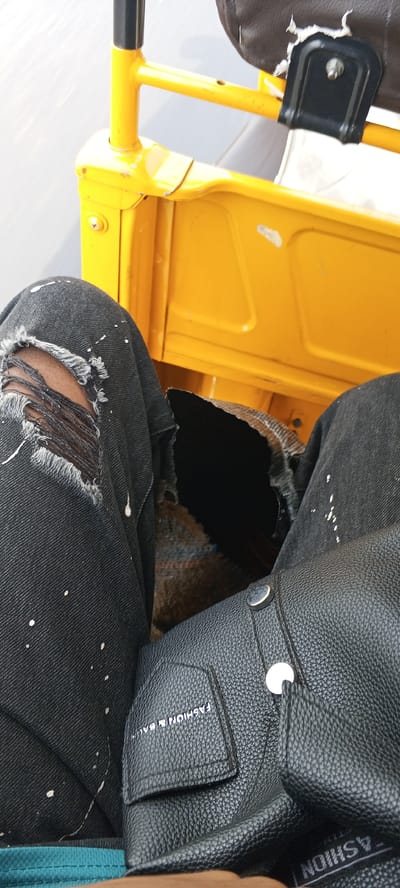 The photo shows a person sitting in a yellow tricycle (Keke Napep) in Uke, Nigeria. The person is wearing ripped black jeans with white paint splatters and has a black textured leather bag resting on their lap. The background includes a portion of the tricycle's yellow metal frame and seat.