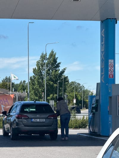 In Tiskre, Estonia, a dark grey Opel Astra station wagon with license plate 252 BXB is parked at a gas station. The car is positioned at a gas pump under a blue canopy that features the ALEXELA logo. A person wearing a grey hooded jacket and blue jeans is standing at the rear of the car, appearing to be operating the fuel pump. The gas station has multiple pumps and is illuminated by bright daylight. The background includes tall green trees, a light pole, and a white flag on a pole with a green logo. Other vehicles and buildings are visible in the distance. The ground is paved, and a white car is partially visible in the foreground. A sign on the gas pump pillar indicates "HADAABI" and the number "112".
