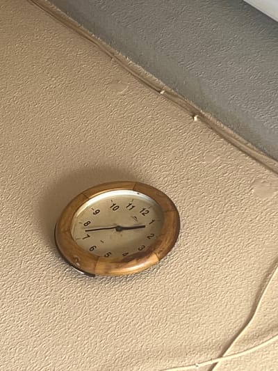 An analog clock with a circular wooden frame is prominently displayed on a textured, light beige wall. The clock face, aged and discolored, features black numeric hour markers from 1 to 12. The hour hand points between 7 and 8, slightly closer to 8, while the minute hand is positioned near the 12, indicating an approximate time of 7:58 or 7:59. A faint shadow is cast beneath the clock, suggesting ambient light from above. In the upper right portion of the image, the light beige wall meets a darker gray, textured surface, possibly a ceiling or an adjacent wall. A light-colored electrical cable runs horizontally along this seam, secured by small white clips. Another similar cable originates from the lower right corner and extends upwards towards the right side of the clock. The entire scene is an indoor environment, located in Ontinyent, Spain. No individuals or actions are depicted.