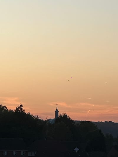 The image, correctly oriented, depicts a serene sunset or sunrise scene over what appears to be a town or village, possibly in High Wycombe, United Kingdom. The sky dominates the frame, showcasing a warm gradient that transitions from a light yellow-cream on the left to soft orange and pink hues towards the right. Several faint, wispy white streaks, likely contrails or thin clouds, are visible against the warmer parts of the sky.
In the upper-middle section of the sky, two very small, brightly colored objects are visible: one appears magenta or pink, and the other is dark, possibly green or black, suggesting they are balloons.
Along the horizon, a dark, silhouetted treeline extends across the lower portion of the frame. To the right, a prominent church steeple, topped with a cross, stands out against the colorful sky. Below the treeline on the far right, the upper part of a dark building with several white-framed windows is partially visible, indicating residential structures. In the distance, behind the treeline and church, the faint silhouette of rolling hills or mountains can be observed.
The time of day is clearly either sunrise or sunset, creating a peaceful and quiet atmosphere. There is no visible text in the image.