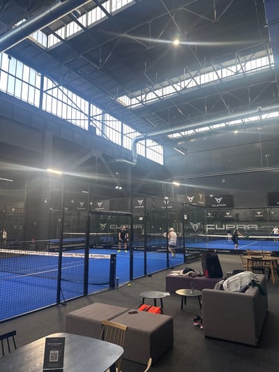 The image shows an indoor padel tennis facility, identified as Padel Up Mülheim, located at Parkhausstraße 2, 45476 Mülheim an der Ruhr, Germany.

The scene captures a large, high-ceilinged industrial-style hall with exposed steel support structures, extensive overhead windows/skylights, and visible silver ventilation ducts. The floor consists of dark gray carpeting in the lounge areas and vibrant blue playing surfaces for the padel courts.

At least three padel courts are visible, enclosed by black mesh fencing and glass walls, each featuring prominent "CUPRA PADEL UP" branding on the back walls and nets. On the nearest court, two adult males are actively playing, one dressed in black and the other in white, both positioned near the net. A yellow padel ball is visible on this court. Further down the hall, additional players are discernible on a second court.

In the foreground and mid-ground, a lounge area is furnished with dark gray cushioned seating modules (ottomans and a sofa), small circular dark-colored side tables, and wooden tables with chairs. Personal effects, including a red and black sports bag, a white duffel bag, red sneakers, and a white towel, are placed on or beside the seating. A single yellow padel ball rests on the carpet in the lounge area. The entire space is brightly illuminated by natural light from the high windows, creating visible lens flares, supplemented by overhead artificial lighting fixtures.