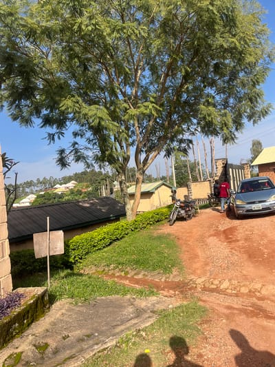The image depicts a dirt road in Gembu, Nigeria, with a car and a motorcycle parked on the side. A person in a red shirt stands near a gate, and another person is partially visible behind the car. Several buildings, including a house with a yellow roof and a larger building with a dark roof, are visible in the background, nestled amongst lush greenery and trees. The sky is clear and blue, indicating a sunny day.
