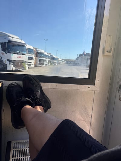 The image shows a person's legs and feet wearing black sneakers and black shorts. The person is sitting inside a cabin, with their legs crossed and resting on a surface. Through a window, a line of parked trucks is visible in an outdoor lot. The sky is clear and blue. The location is identified as Kapitan Andreevo, Bulgaria. The environment appears to be a rest stop or parking area for trucks.
