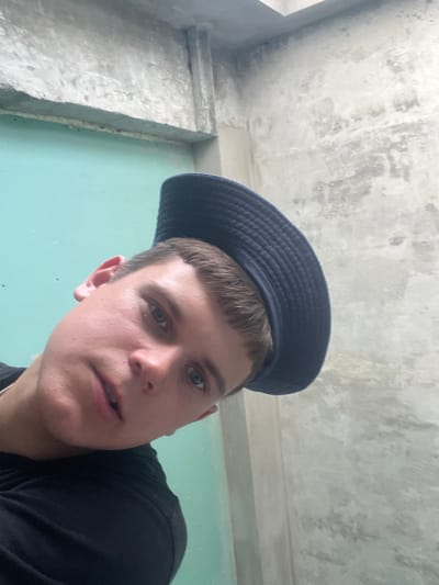 A young person, with fair skin, short brown hair, and blue eyes, is looking up at the camera. They are wearing a dark blue hat with a textured surface and a black t-shirt. The hat is positioned low on their forehead, partially obscuring their eyebrows. The person's expression appears neutral, with their mouth slightly open.
The background consists of a rough, textured concrete ceiling with visible imperfections and stains, suggesting an unfinished or industrial interior. To the left, a light-colored concrete structure with sharp edges is visible, possibly a wall or support beam. The right side of the background is a muted teal color, possibly a wall or painted surface. The lighting suggests an indoor environment with diffused light, making it difficult to determine the time of day. There are no visible signs of weather conditions.
The perspective of the photo is from below, looking up at the subject, creating a sense of being looked down upon. This low-angle shot, combined with the subject's upward gaze, gives the image a direct and engaging feel. The overall impression is that of a casual, perhaps candid, self-portrait taken in an unadorned, possibly rural or construction-related setting within Аснежыцкі сельскі Савет, Belarus. No text is visible in the image.