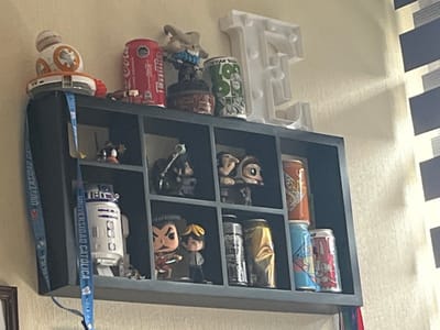 The image displays a dark grey, multi-compartment wall shelf mounted on a light-colored, textured wall. On top of the shelf, from left to right, are a white and orange Star Wars BB-8 droid figure on a base, a red Coca-Cola soda can, a small figurine on a dark base, a green patterned metal can, and a white, light-up decorative letter 'E'. The letter 'E' casts a subtle shadow on the wall.
Inside the shelf's top row, from left to right, are a small figurine with orange accents, a dark-colored action figure, two distinct small figurines (one with a light brown head), and an orange beverage can. The bottom row of compartments, from left to right, contains a white and blue Star Wars R2-D2 droid figure, two "Funko Pop!" style figures (one red-bodied, one grey-bodied with yellow hair), three metallic beverage cans (one dark, one crumpled gold/yellow, one light blue with colorful graphics), and a white beverage can with elaborate colorful artwork.
A blue lanyard, prominently displaying "UNIVERSIDAD CATOLICA" and "VOLLEYS" in white text, hangs from the left side of the shelf. A portion of a framed item is visible in the bottom left corner, beneath the lanyard. To the right of the shelf, a window with dark and light horizontal blinds is partially visible. The scene is an interior room, likely a personal space, in La Serena, Chile, with the "UNIVERSIDAD CATOLICA" lanyard indicating an affiliation with the Universidad Católica del Norte campus serving the La Serena-Coquimbo conurbation.
