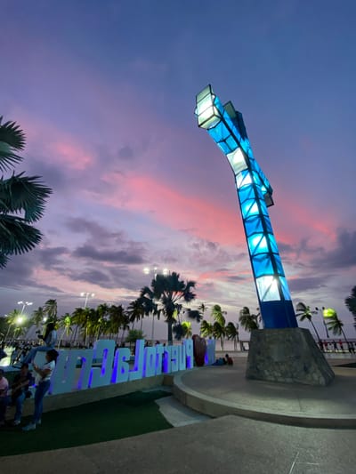 The image shows a street scene in Puerto La Cruz, Venezuela, under a colorful twilight sky. A large, illuminated blue and white structure stands prominently, possibly a decorative archway. Along the sidewalk, illuminated blue letters spell out "Puerto La Cruz." Palm trees line the street. People are visible near the illuminated letters, including some who appear to be posing or taking pictures. The ground consists of concrete and a patch of grass. A stone structure is visible near the base of the archway. The sky is a blend of blues, purples, and pinks with cloud cover. Streetlights are also visible along the road.