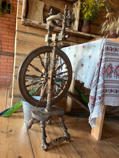 A close-up, vertically oriented medium shot captures a spinning wheel and a piece of embroidered fabric in a rustic, indoor setting. The spinning wheel, made of aged, dark wood, is the central object, with its large wheel and intricate details visible. To the upper right of the spinning wheel, a light-colored fabric with red and grey embroidered floral patterns is draped over a wooden structure, suggesting a workshop or display area.

In the background, wooden plank walls and shelves are visible, adding to the rustic ambiance. On a shelf to the upper left, a clay jug sits next to a bouquet of dried wheat and yellow wildflowers, hinting at a rural or traditional setting. A patterned, light-colored object with intricate carved details is placed on a wooden shelf below the spinning wheel. A galvanized metal bucket is also visible near the base of the spinning wheel.

The overall impression is one of traditional craftsmanship and a connection to rural life in Russia, possibly in Vybolovo given the location context. The lighting appears to be natural, coming from an unseen source, and the overall mood is one of quiet history and handmade artistry. There is no visible text in the image.