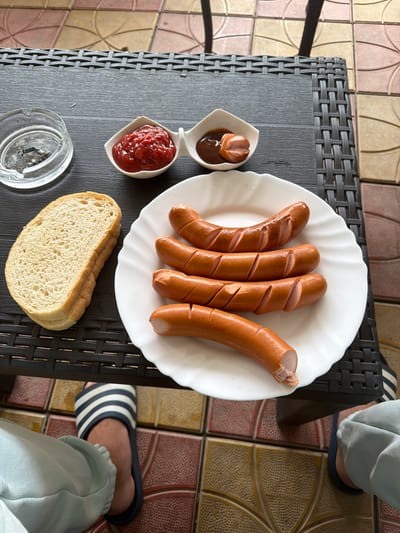 An overhead shot captures a simple meal laid out on a dark grey, woven-patterned table, likely on an outdoor patio or balcony in Dragor, Bulgaria. The main focus is a white scalloped plate holding five cooked hot dogs or sausages. Four of the sausages exhibit diagonal cuts along their length, while one on the far right has a bite taken from its end, revealing the light pink interior.
Adjacent to the plate are two small white, heart-shaped dishes containing condiments. One dish holds a dollop of red ketchup, and the other contains a darker, brown sauce (possibly BBQ sauce or mustard) with a small, cut piece of sausage submerged within it. A thick slice of white bread rests on the table next to the condiments.
In the bottom left corner of the frame, a clear glass ashtray with some ash is visible. The lower right portion of the image shows the legs and feet of a person, dressed in light blue casual pants and wearing black-and-white striped flip-flops or sandals. The floor beneath the table is adorned with decorative, multi-toned tiles featuring geometric patterns in shades of brown, beige, and pink. The lighting is bright and even, suggesting daytime. No visible text is present in the image.