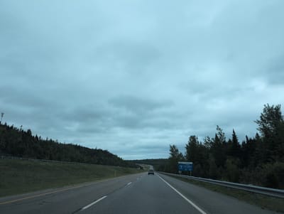 The image captures a view from a vehicle traveling on a highway, likely in or near the Town of Hampton, Canada. The sky is overcast with a significant amount of cloud cover, casting a muted light over the scene. A two-lane highway stretches forward, with a white line marking the center and a yellow line to the left. A silver sedan is visible ahead, traveling in the same direction.

On the left side of the highway, a grassy embankment slopes upwards, topped by a dense forest of evergreen trees. A guardrail runs along the edge of the embankment. On the right side of the highway, another dense forest of evergreen trees lines the road. A blue highway sign is positioned on the right, indicating "Hampton Visitor Information aux visiteurs" and listing "EXIT 158 ROUTE 1". A question mark symbol is also visible on the sign. The overall environment suggests a rural or semi-rural highway setting, possibly within a forested area of Canada.