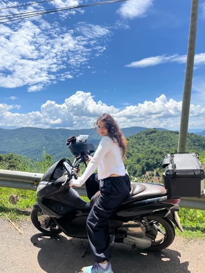 A young woman with curly brown hair sits on a black scooter on a road in Son Dien, Vietnam. She is wearing a white long-sleeved shirt and dark pants. The scooter has a black helmet on its handlebars and a black storage box on the back. The woman is looking over her shoulder at the camera. In the background, there are lush green mountains covered in trees, and a blue sky with white, puffy clouds. A metal guardrail runs along the side of the road, and a utility pole is visible on the right side of the image. The ground is paved with asphalt, and there is some grass and vegetation on the roadside.