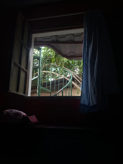An open window reveals a green spiral staircase surrounded by foliage, with a blue curtain partially obscuring the view. Two pillows are visible on a dark surface in the foreground. The scene is dimly lit, suggesting an indoor setting at dusk or in a shaded area. The image appears to be taken from within a room in Managua, Nicaragua, though no specific landmarks are identifiable.