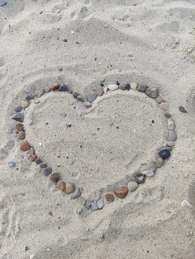 A heart shape is formed by a line of small, rounded pebbles on a sandy surface. The pebbles vary in color from light gray to dark gray and brown, with some appearing speckled. The sand is a light tan color and shows slight undulations and indentations, suggesting it is beach sand. The image is taken from a top-down perspective. The location in the city of Nezavertaylovka, Moldova is not discernible from the visual information in the image. There are no people or any other objects present in the scene.