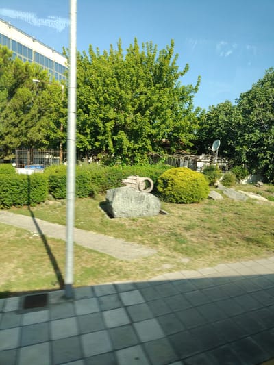 The image captures a sunlit outdoor scene, likely in Larissa, Greece, showcasing a blend of urban and natural elements. In the background, a modern building with a light-colored facade and numerous windows stands tall. Lush green trees, including a large, dense one, dominate the mid-ground. To the right, a satellite dish is visible, mounted on a pole. In the foreground, a grey, textured paved area transitions to a grassy area with a pathway. A large, grey boulder with a circular metallic object resting on it is a prominent feature in the middle ground. A tall, metallic pole stands vertically in the center of the image, casting a long shadow. The sky is a clear, bright blue. The overall impression is a clean and orderly environment, possibly a public space or park adjacent to a building.